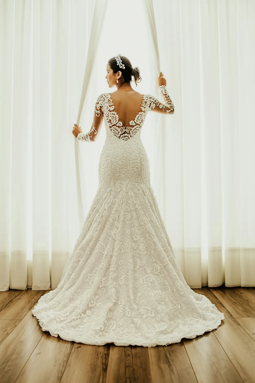 A bride in a fitted lace gown opening sheer curtains, seen from behind