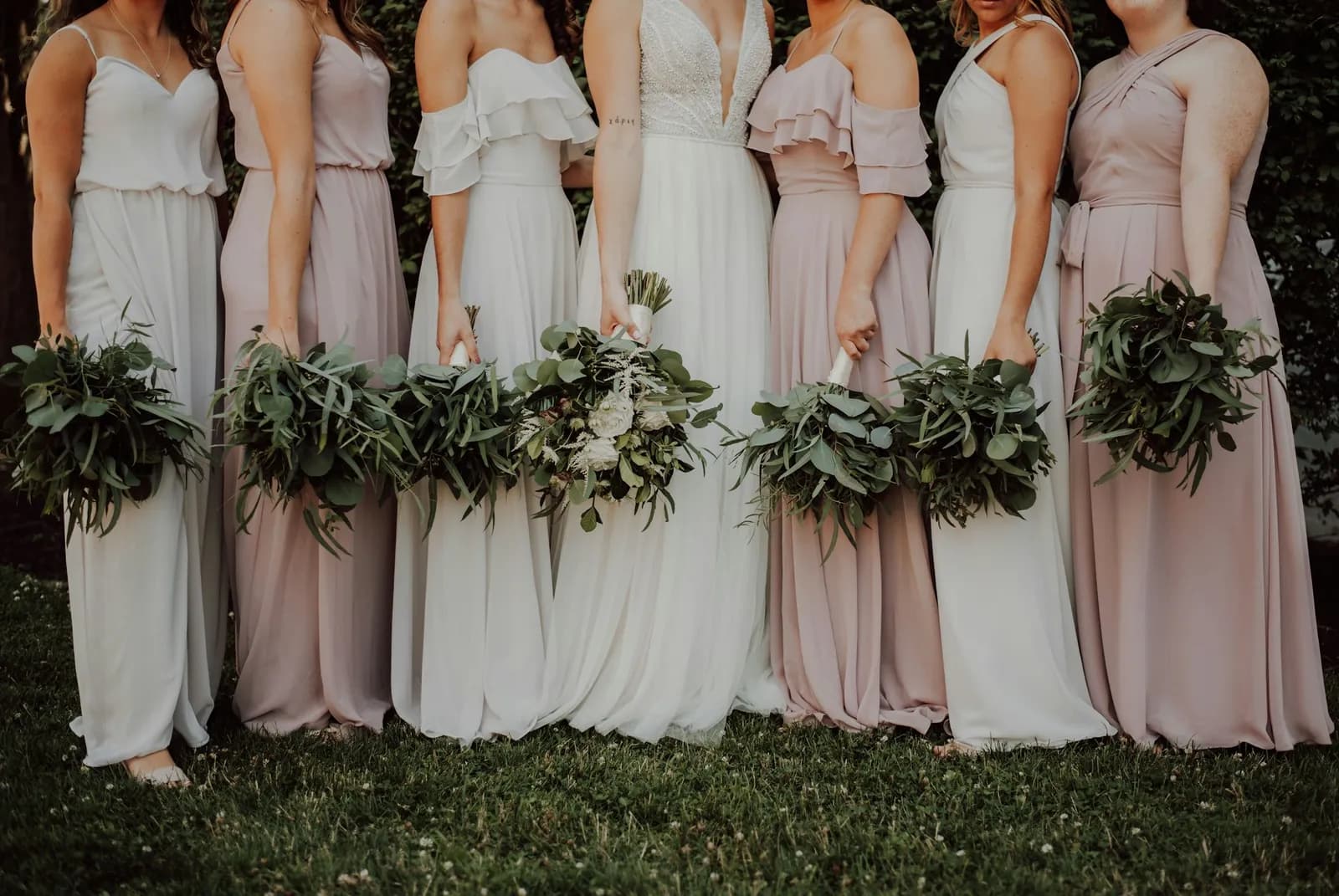 A row of bridesmaids in blush and ivory dresses holding greenery bouquets